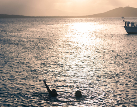 Two people swimming in a lake at sunset with a boat nearby.