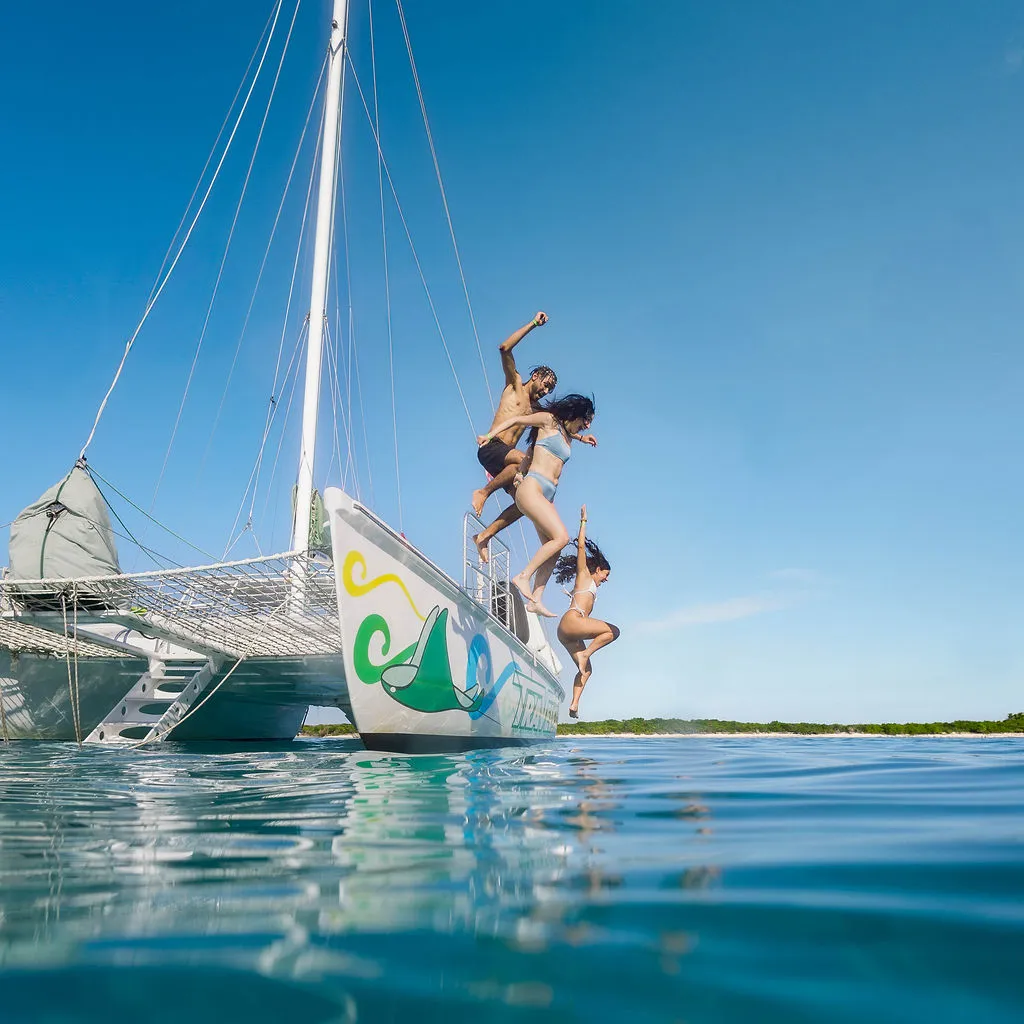 Three people jumping off a sailboat into the ocean under a clear sky.
