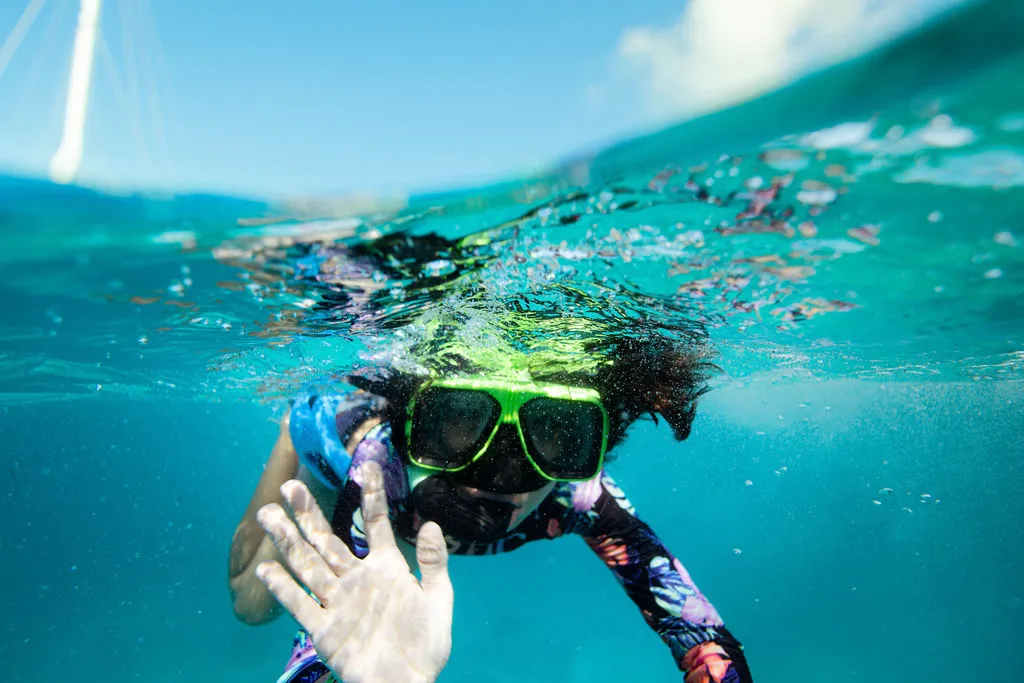 Person snorkeling underwater, wearing a colorful wetsuit and green goggles, waving at the camera.